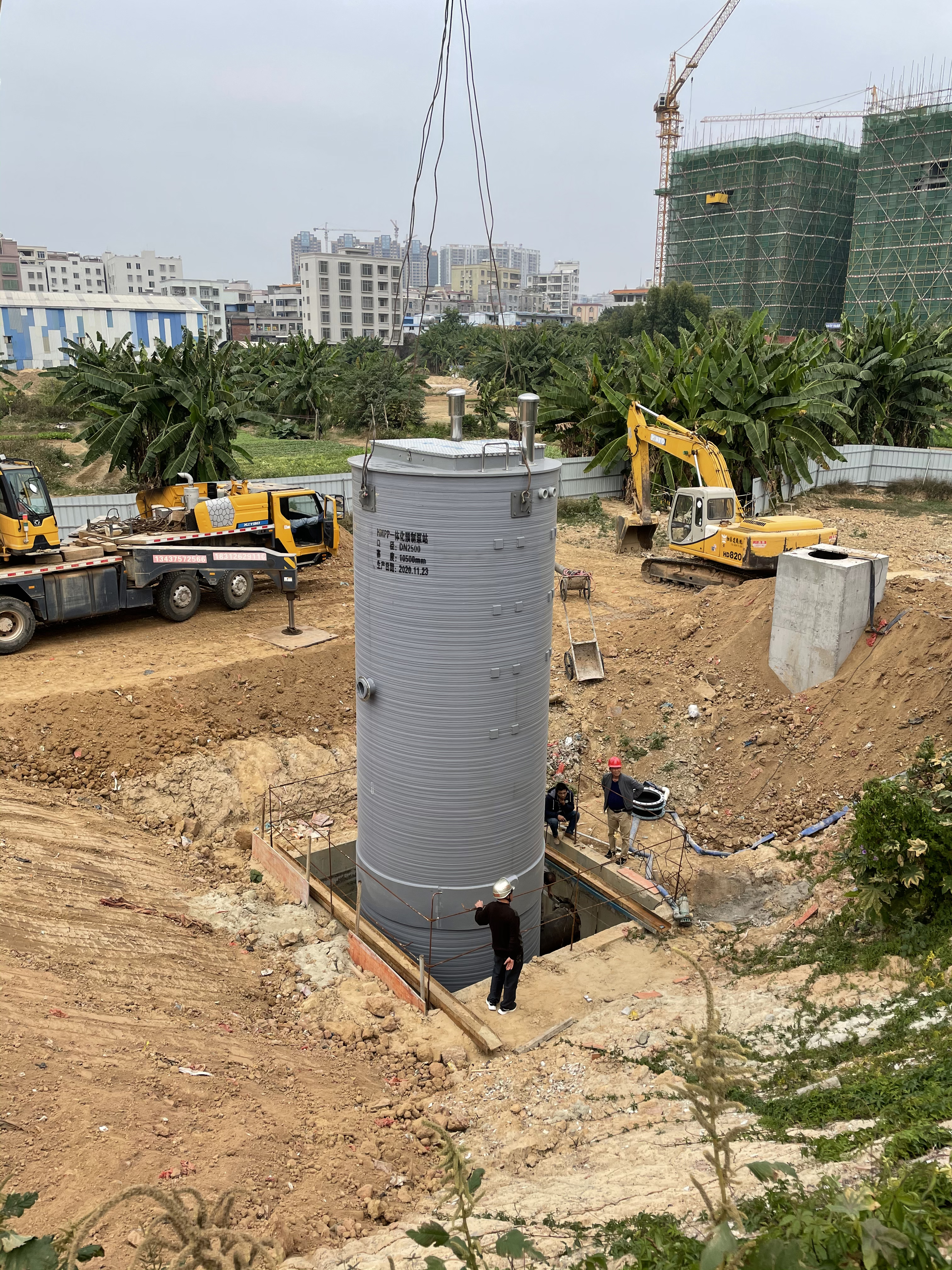 Prefabricated pumping station lifted by crane into an excavation at a construction site, with workers beside the pit